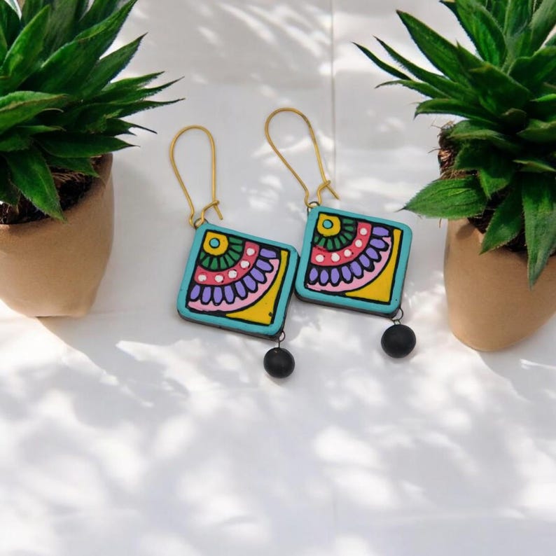 Puede incluir: Un par de pendientes cuadrados de color azul turquesa con dise&ntilde;os florales coloridos y detalles de cuentas negras. Los pendientes tienen ganchos dorados.