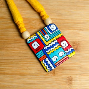Puede incluir: Un collar con colgante geom&eacute;trico y colorido. El colgante es un rect&aacute;ngulo con patrones rojos, azules, amarillos y blancos. El collar tiene un cord&oacute;n amarillo y cuentas de madera. El fondo es una superficie de madera.