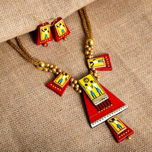 May include: A red and yellow terracotta jewelry set with a necklace and earrings. The necklace features a large triangular pendant with a smaller rectangular pendant attached. Both pendants have painted figures and gold accents. The earrings are small and rectangular with painted figures.