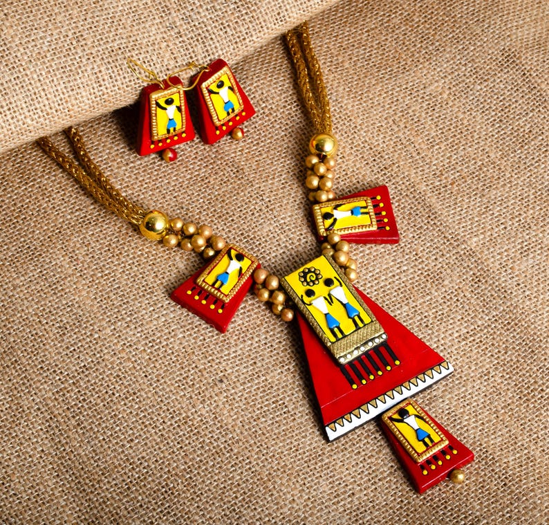 May include: A red and yellow terracotta jewelry set with a necklace and earrings. The necklace features a large triangular pendant with a smaller rectangular pendant attached. Both pendants have painted figures and gold accents. The earrings are small and rectangular with painted figures.