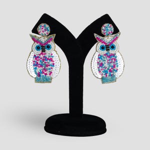 May include: A pair of owl-shaped earrings with pink, blue and white sequins and beads. The earrings are displayed on a black stand.