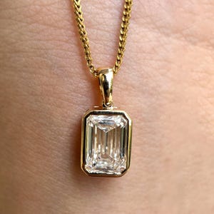 Puede incluir: Un collar con colgante de oro con una piedra preciosa rectangular y transparente. La piedra está engastada en un bisel de oro y cuelga de una fina cadena de oro. El colgante es una joya clásica y elegante.