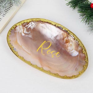 Könnte beinhalten: Eine ovale Schale aus Perlmutt mit einem goldfarbenen, verzierten Rand. Das Wort "Rose" ist in goldener Schrift auf die Schale geschrieben. Dekorative Grünpflanzen und ein Buch befinden sich im Hintergrund.