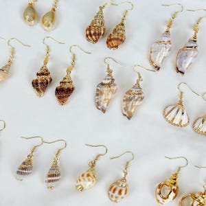 May include: A collection of seashell earrings with gold-colored hooks. The earrings feature various shell shapes and colors, including white, brown, and tan. Some earrings have gold accents.