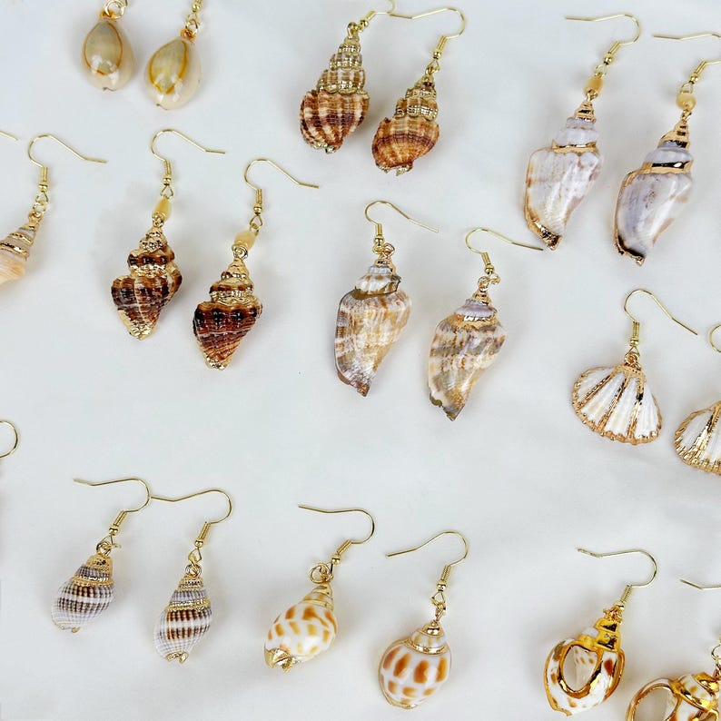 May include: A collection of seashell earrings with gold-colored hooks. The earrings feature various shell shapes and colors, including white, brown, and tan. Some earrings have gold accents.