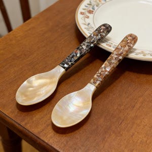 Può includere: Due cucchiai decorativi con conche di madreperla e manici a mosaico. Un manico è nero e l'altro marrone. I cucchiai sono su una superficie di legno vicino a un piatto con motivi floreali.