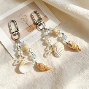 Könnte beinhalten: Zwei Schlüsselanhänger mit silbernen Verschlüssen, verziert mit Muscheln, Kunstperlen, Holzperlen und transparenten sternförmigen Anhängern. Ein Schlüsselanhänger hat eine weiße Muschel, der andere eine braune und weiße Muschel. Auf neutralem Hintergrund.