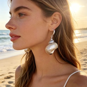 Könnte beinhalten: Silberne Ohrringe mit einem natürlichen Muscheldesign und Perlmutt-Finish. Die Ohrringe hängen an einem silbernen Bügel. Ein einzigartiges Accessoire für Schmuck im Strand- oder Ozean-Stil.
