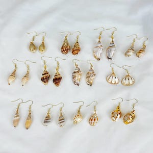 May include: A collection of seashell earrings with gold-colored hooks. The earrings feature various natural shell shapes and colors, including white, brown, and tan. Some shells have a spiral design, while others are smooth or textured.