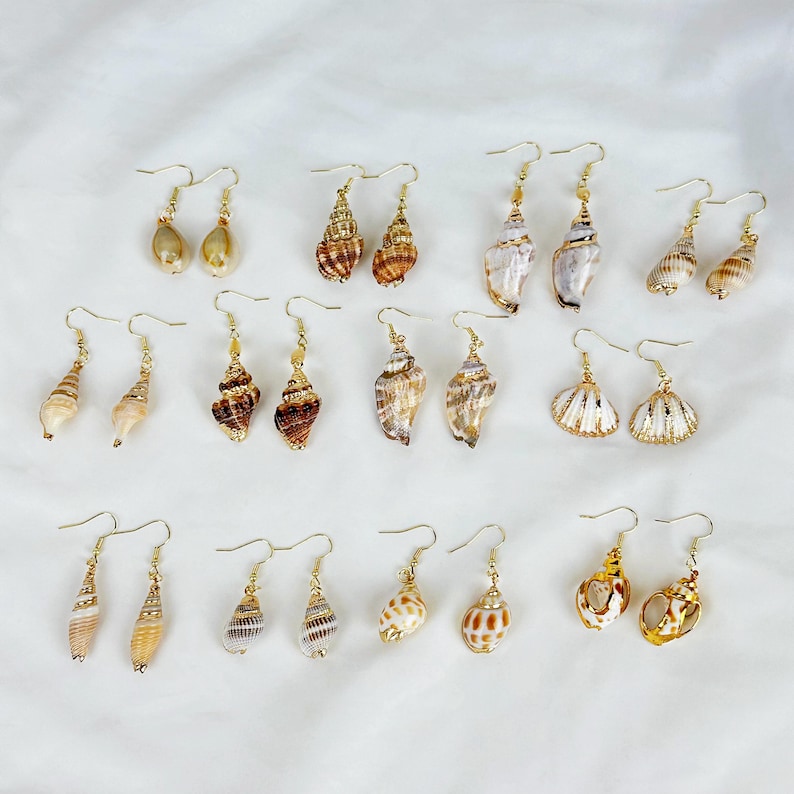 May include: A collection of seashell earrings with gold-colored hooks. The earrings feature various natural shell shapes and colors, including white, brown, and tan. Some shells have a spiral design, while others are smooth or textured.