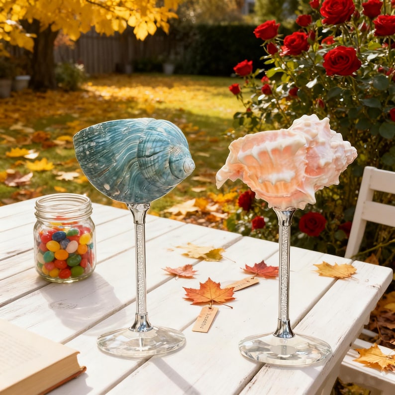 May include: Two decorative wine glasses with seashell bowls, one teal and one peach, sit on a white table. The stems are embellished with crystals. A jar of jelly beans and autumn leaves are also on the table, with a backdrop of a garden.