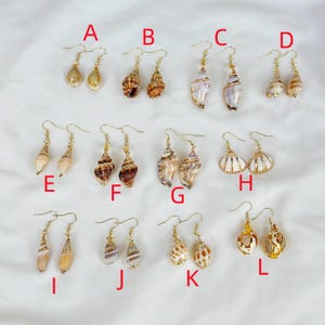 May include: A collection of twelve pairs of seashell earrings, each with a unique design. The earrings feature various shell shapes and colors, with gold-colored hooks. The earrings are arranged on a white surface.