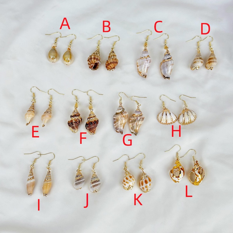 May include: A collection of twelve pairs of seashell earrings, each with a unique design. The earrings feature various shell shapes and colors, with gold-colored hooks. The earrings are arranged on a white surface.