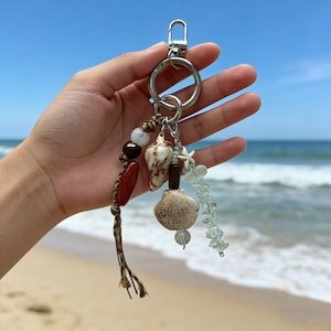 May include: A beach-themed keychain with a silver clasp, featuring seashells, beads, and a small starfish charm. The keychain includes a variety of natural elements in shades of brown, white, and clear, creating a coastal aesthetic.
