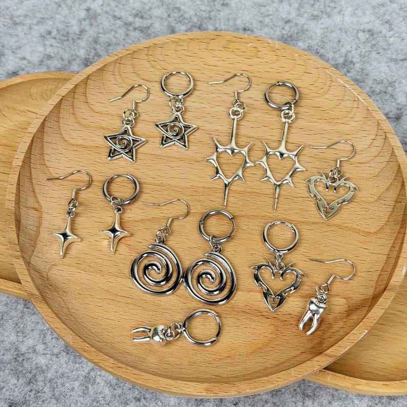 May include: A collection of silver-toned earrings displayed on a wooden tray. The earrings feature various designs, including stars, spirals, hearts, and a tooth. Some earrings have a hook closure, while others have a hoop.