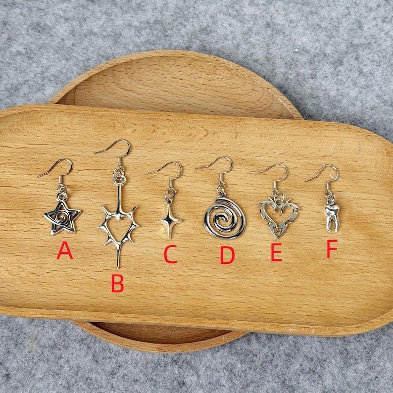 May include: A collection of six silver-toned earrings displayed on a wooden tray. The earrings feature various designs, including a star, a heart, a spiral, a tooth, and a starburst. Each earring has a hook for pierced ears.