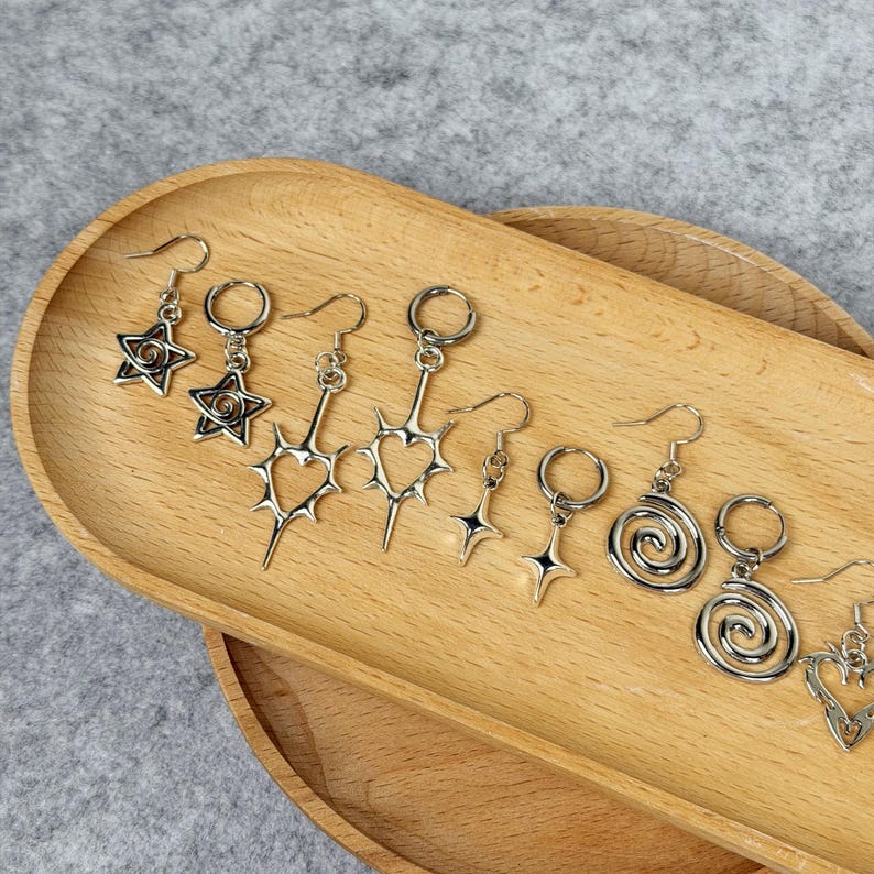 May include: A collection of silver-toned earrings displayed on a wooden tray. The earrings feature various designs, including stars, hearts, and spiral shapes. Each earring has a hook for easy wear.
