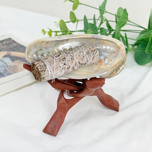 Könnte beinhalten: Abalone-Muschelschale mit einem weißen Salbei-Bündel, das auf einem geschnitzten Holzständer ruht. Die Muschel hat schillernde Farben, und der Salbei ist mit weißer Schnur gebunden. Der Ständer ist dunkelrotbraun.