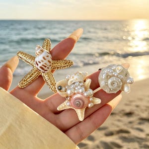 Op de afbeelding: Drie schelpenringen worden getoond tegen een strandachtergrond. Eén ring heeft een gouden zeester met een schelp, een andere heeft een zeester met parels en een schelp, en de derde heeft een schelp met parels.