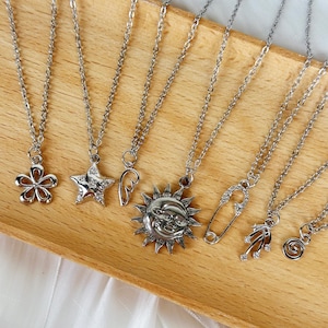 Könnte beinhalten: Sieben silberne Halsketten mit verschiedenen Anhängern, darunter eine Blume, ein Stern, ein Herz, eine Sonne, eine Sicherheitsnadel und zwei abstrakte Designs. Die Halsketten werden auf einer Holzoberfläche präsentiert.
