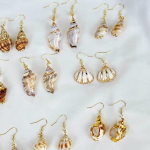May include: A collection of seashell earrings with gold-colored hooks. The earrings feature various shell shapes and colors, including white, brown, and tan. Some earrings have gold accents. The earrings are displayed on a white surface.