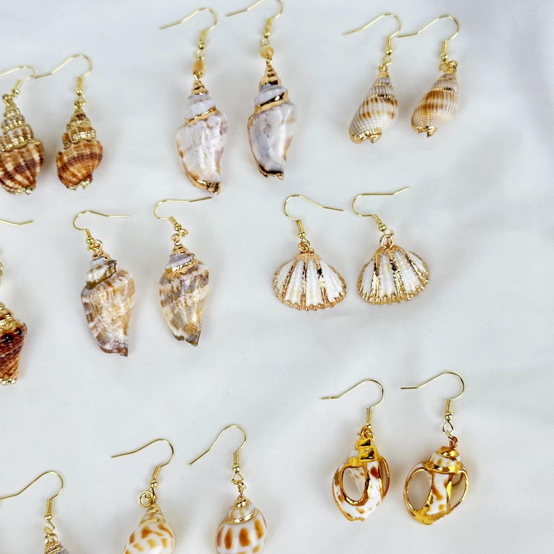 May include: A collection of seashell earrings with gold-colored hooks. The earrings feature various shell shapes and colors, including white, brown, and tan. Some earrings have gold accents. The earrings are displayed on a white surface.