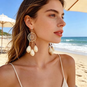 Könnte beinhalten: Goldfarbene Traumfänger-Ohrringe mit kreisförmigem Netzdesign und herabhängenden tropfenförmigen Muscheln. Die Ohrringe haben auch kleine braune Perlen. Getragen von einer Person am Strand.