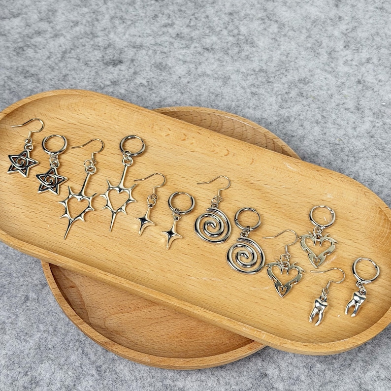 May include: A collection of silver-toned earrings displayed on a wooden tray. The earrings feature various designs, including stars, hearts, spirals, and teeth. The earrings are designed with a hook closure.