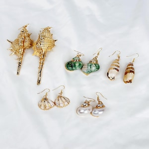 Peut inclure: Une collection de boucles d'oreilles coquillages de différents modèles. Les boucles d'oreilles présentent des accents dorés et sont dans des tons de blanc, de vert et de marron. Les boucles d'oreilles sont présentées sur une surface blanche.