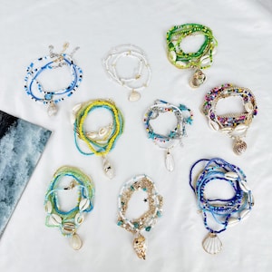 Può includere: Una collezione di collane e braccialetti di perline in vari colori, tra cui blu, bianco, giallo e verde. Ogni pezzo presenta conchiglie e ciondoli. I gioielli sono disposti su una superficie bianca, mostrando un tema costiero.