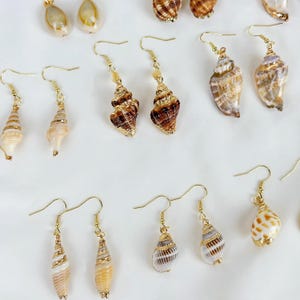 May include: A collection of seashell earrings with gold-colored hooks. The earrings feature various shell shapes and colors, including tan, brown, and white. Some earrings have small beads. The earrings are displayed on a white surface.