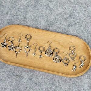 May include: A collection of silver-toned earrings with various designs, including stars, hearts, spirals, and teeth, displayed on a wooden tray. The earrings have a gothic or alternative aesthetic, with intricate details and a polished finish.