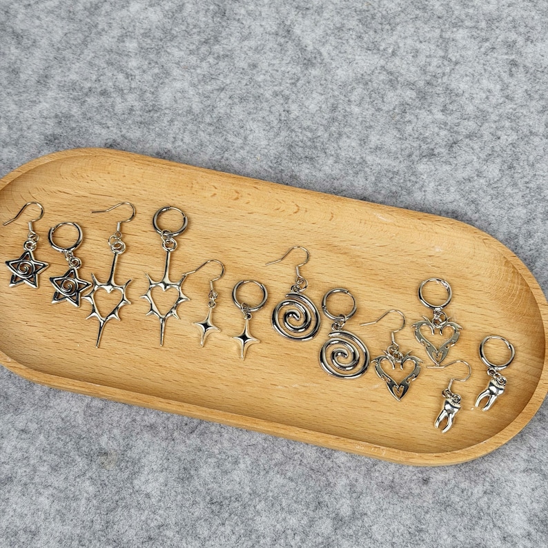 May include: A collection of silver-toned earrings with various designs, including stars, hearts, spirals, and teeth, displayed on a wooden tray. The earrings have a gothic or alternative aesthetic, with intricate details and a polished finish.