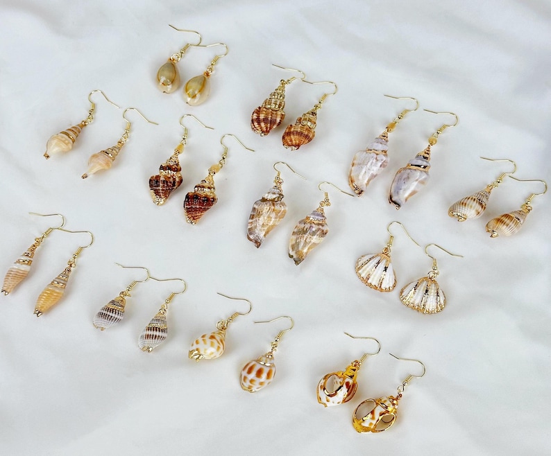 May include: A collection of seashell earrings with gold-colored hooks. The earrings feature various shell shapes and colors, including white, brown, and tan. The earrings are displayed on a white surface.