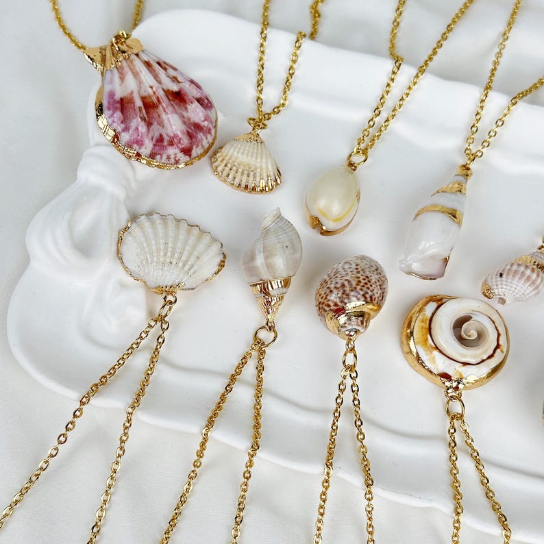 Op de afbeelding: Een verzameling gouden kettingen met verschillende zeeschelpen. De schelpen hebben verschillende vormen en kleuren, waaronder wit, bruin en roze. Elke schelp is bevestigd aan een fijne gouden ketting, wat een sieradenset in strandstijl cre&euml;ert.