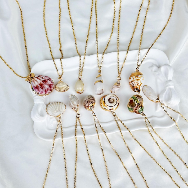 Op de afbeelding: Een verzameling goudkleurige kettingen met verschillende schelpen als hangers. De schelpen vertonen een reeks kleuren, waaronder wit, roze, bruin en goud. De kettingen zijn gerangschikt op een wit dienblad tegen een witte achtergrond.