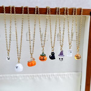 Op de afbeelding: Een verzameling goudkleurige kettingkettingen, elk versierd met een Halloween-thema hanger. Hangers zijn een spook, pompoen, zwarte kat en heksenhoed. De kettingen worden gepresenteerd in een houten sieradendoos.