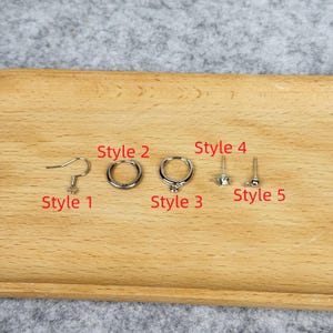 May include: A collection of five different styles of silver-colored earrings displayed on a wooden surface. Styles include a fish hook, hoop, ring with a gem, and two stud earrings. The earrings are arranged in a row.