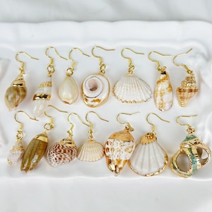 Peut inclure: Une collection de boucles d'oreilles en coquillages avec des crochets dorés. Les boucles d'oreilles présentent diverses formes et tailles de coquillages, certaines ornées de détails dorés. Les coquillages affichent des couleurs naturelles, blanc, beige et marron.