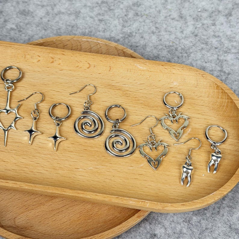May include: A collection of silver-toned earrings with various designs, including hearts, stars, spirals, and teeth, displayed on a wooden tray. The earrings have different hook styles and sizes, showcasing a variety of aesthetic choices.