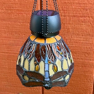 May include: A hanging solar-powered lamp featuring a stained glass shade with a dragonfly motif. The shade is constructed from amber, cream, and brown glass pieces joined by dark metal solder. A black solar panel cap sits atop the shade.