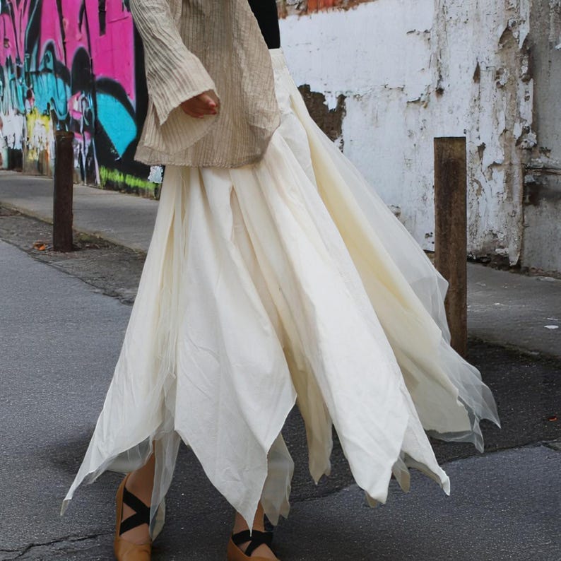 May include: A cream-colored, flowing skirt with a unique petal-like hemline. The skirt is paired with a beige top with flared sleeves. The outfit is completed with brown flats with black straps. The background features a graffiti-covered wall.