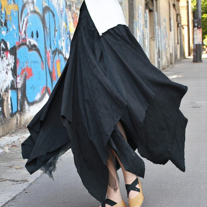 May include: A flowing black skirt with an asymmetrical hemline. The skirt is layered and appears to be made of a lightweight fabric. The model is wearing black shoes with a cross-strap design. The background features a graffiti-covered wall.