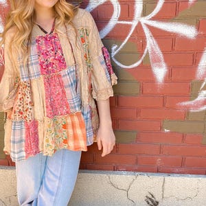 May include: A patchwork top in pink, beige, and orange, featuring floral, plaid, and solid panels. The top has a loose fit, ruffled sleeves, and a square neckline. The model is wearing light blue jeans. The top is made of various fabrics.
