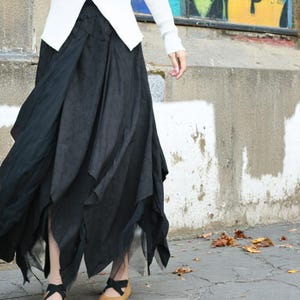 May include: A black, asymmetrical skirt with a layered design. The skirt has a flowing, textured appearance, with a longer length and a unique, irregular hemline. The model is wearing a white sweater and tan shoes.