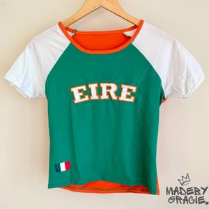 Può includere: Una maglietta verde, bianca e arancione con la parola "EIRE" in lettere bianche con contorno arancione. La maglietta ha un bordo bianco e arancione intorno al collo e una piccola toppa della bandiera irlandese sul lato sinistro.