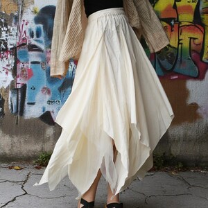 May include: A cream-colored, asymmetrical skirt with a layered design. The skirt has a flowing, lightweight fabric and a unique cut that creates a dynamic silhouette. The model is wearing black sandals. The background is a graffiti-covered wall.