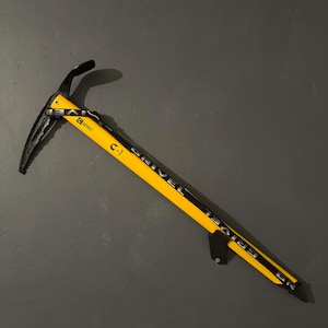 May include: A yellow and black ice axe with the text "GRIVEL" and "C-1" printed on the shaft. The ice axe has a curved pick and adze, and is designed for climbing. The axe is mounted on a dark gray surface.