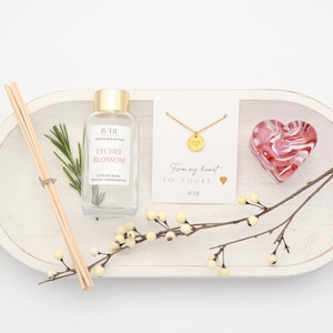 May include: A white wooden tray displays a selection of gifts. A bottle of Lychee Blossom fragrance, a gold heart pendant necklace, and a heart-shaped ornament are arranged with sprigs and a reed diffuser.