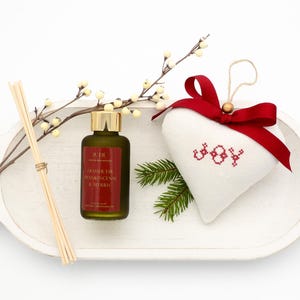 Könnte beinhalten: Festliche Anordnung mit einer grünen Flasche, einem goldenen Deckel und einem weißen herzförmigen Ornament mit rotem "JOY"-Stickerei. Ein rotes Band, ein Beeren-Zweig, ein Kiefernzweig und ein Schilfrohrdiffusor sind ebenfalls enthalten.